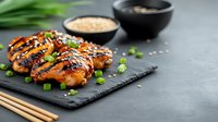 Grilled teriyaki chicken wings garnished with sesame seeds and green onions served on black slate with dipping sauces and chopsticks against dark background.