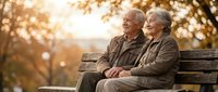 Happy senior Caucasian couple sitting together on park bench during golden hour sunset, enjoying peaceful retirement moment outdoors in autumn nature.