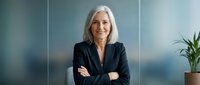 Mature woman with white hair wearing black blazer, arms crossed, smiling at camera in modern office setting with plant and blurred background. Corporate portrait.
