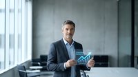 Confident mature Caucasian businessman in suit holding digital tablet displaying financial charts and graphs in modern office conference room.