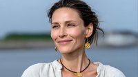 Confident middle-aged woman with gold earrings smiling outdoors against blurred waterfront background, wearing casual white top and necklace.