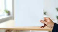 Person with medium skin tone holding blank white paper sheet in bright interior space with natural window light and blurred green plants, suitable for mockups and presentations.