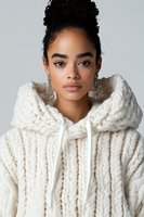 Young African American woman wearing chunky knit white hoodie sweater and crystal earrings poses against light background for fashion and beauty portrait.