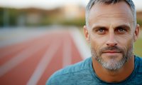 Confident mature Caucasian male athlete with gray beard wearing blue sportswear standing on running track during outdoor training session at stadium.