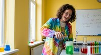 Young mixed-race female student conducting colorful chemistry experiment in bright yellow laboratory classroom wearing tie-dye shirt with beakers and solutions.