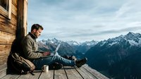 Young man working remotely on laptop while sitting on wooden mountain cabin terrace with stunning alpine peaks backdrop for digital nomad lifestyle.