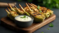 Grilled artichoke halves with creamy dipping sauce served on wooden board. Fresh herbs garnish and bamboo skewers create elegant appetizer presentation.