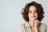 Young woman with curly brown hair and natural makeup smiling confidently while resting chin on hand against light background, wearing beige blazer.