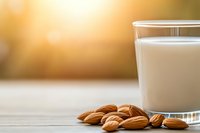 Fresh glass of almond milk with raw almonds scattered on wooden table surface against warm bokeh background, healthy dairy alternative beverage.