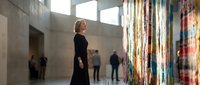 Young blonde woman in black dress contemplating colorful abstract art installation in modern concrete gallery space with natural lighting.