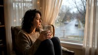 Young Caucasian woman in cozy sweater sitting in armchair with hot coffee cup, gazing through rainy window at home in thoughtful mood.