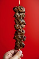 Grilled meat kebab skewer held by hand against vibrant red background, juicy marinated chunks showing char marks and caramelization for food photography.