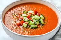 Fresh gazpacho soup garnished with diced cucumber and tomatoes in white bowl, drizzled with olive oil. Bright orange-red color creates appetizing presentation.