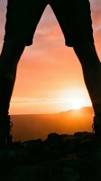 Silhouette of hiker legs framing golden sunrise through mountain cave opening with warm orange sky and distant peaks for adventure travel concepts.