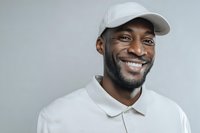 Happy African American man in white baseball cap and polo shirt smiling warmly against gray background for lifestyle and portrait photography.