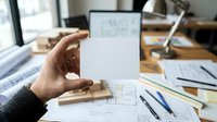 Young male professional holding blank white card at wooden desk with architectural blueprints, laptop, and design tools for branding, business card mockup, or corporate identity presentation.