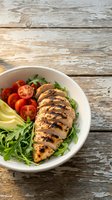 Grilled chicken breast sliced and arranged on fresh arugula with tomato wedges and avocado in white bowl on rustic wooden surface for healthy meal prep and nutrition content.