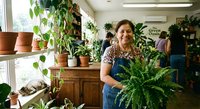 Happy middle-aged Caucasian woman holding large fern plant in bright greenhouse nursery surrounded by potted plants and greenery for gardening business.