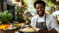 Young African American male chef in white uniform and gray apron presenting freshly prepared creamy risotto dish with herbs and vegetables in restaurant kitchen setting.