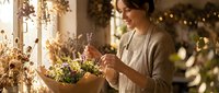 Young woman florist arranging dried flower bouquet in cozy workshop with warm lighting and natural decorations for small business owner.