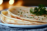 Fresh homemade tortillas stacked on white plate, garnished with herbs and spices, warm golden brown spots on surface, soft texture visible in natural light.