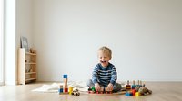 Young blonde boy, approximately 3-5 years old, Caucasian ethnicity, engaged in educational play with colorful wooden blocks and toys on light wooden floor in bright, minimalist interior space.