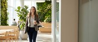 Young professional businesswoman walking in modern eco-friendly office with green plants, holding coffee cup and laptop bag during morning commute.