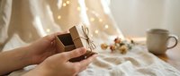 Hands holding small wrapped gift box with twine bow on cozy bed with warm fairy lights creating intimate holiday morning atmosphere for gifting celebrations.