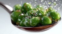 Fresh green peas on metal spoon with water drops splashing, macro food photography showing vibrant vegetables in dynamic composition for culinary design.