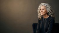 Mature woman with long gray curly hair wearing dark sweater, seated against neutral brown background, professional portrait for business, corporate, and lifestyle design projects.