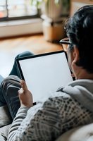 Asian man in casual wear relaxing on couch while using digital tablet with blank white screen, view from behind. Suitable for technology and lifestyle ads.