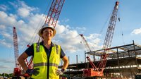Young Asian female construction worker in safety vest and hard hat smiling confidently at building site with cranes and blue sky background.