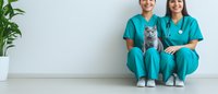 Two veterinary professionals in turquoise scrubs sitting on floor with British shorthair cat against white wall, green plant in corner, modern medical setting.