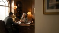 Senior woman with gray hair and middle-aged man in business meeting at wooden desk in elegant office with warm lighting, suitable for corporate, professional, and business communication concepts.