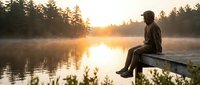 Man in casual clothing sitting on wooden dock at misty lake during golden hour sunrise or sunset, contemplating nature and water reflections for wellness and outdoor lifestyle content.