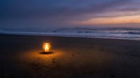 Glowing lantern on sandy beach at twilight with ocean waves and purple sunset sky creating romantic coastal atmosphere for relaxation and meditation.