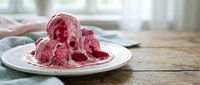 Homemade pink raspberry mousse or whipped cream dessert served on white plate with wooden table and blurred kitchen background, suitable for food blogs and culinary content.