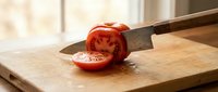 Ripe tomato being sliced with sharp knife on wooden cutting board in bright kitchen, food preparation and cooking ingredient.