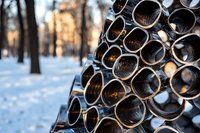 Stack of metal pipes or tubes arranged in geometric pattern against snowy winter forest background with soft bokeh sunset light creating industrial contrast.