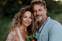 Mature Caucasian couple embracing outdoors, sharing intimate moment. Woman with curly blonde hair in lace dress, man with gray beard wearing denim shirt, natural lighting.