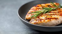 Grilled chicken breast with crispy grill marks and fresh rosemary sprig served on gray ceramic plate, close-up view against blurred background. Food photography.