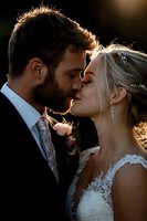 Romantic sunset silhouette of newlyweds sharing intimate kiss. Bride in lace dress with crystal accessories and groom with beard in suit and floral tie.
