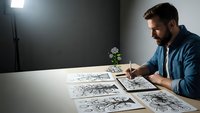 Young bearded male artist drawing detailed sketches at desk in studio with professional lighting setup and small potted plant.