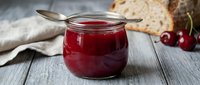 Homemade cherry jam in glass jar with spoon, fresh berries and bread on wooden table for breakfast or baking.