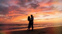 Romantic couple silhouette embracing on beach at sunset with dramatic orange and pink sky clouds reflecting over ocean waves during golden hour.