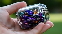 Colorful glass marbles in transparent jar held in palm against blurred green background, showcasing iridescent purple, blue and amber colors with light reflections.
