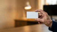 Senior man with wrinkled hands holding blank white business card in modern interior space with warm lighting, suitable for corporate branding and business presentations.