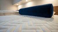 Dark blue cylindrical bolster pillow resting on beige geometric patterned bedding in modern bedroom with warm lighting and wooden headboard.