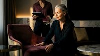 Mature Caucasian businesswoman working on laptop in modern office lounge while colleague reads documents in background during evening hours.