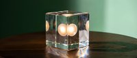 Glowing candles inside transparent cube on wooden surface against green background, modern home decor and ambient lighting design element.